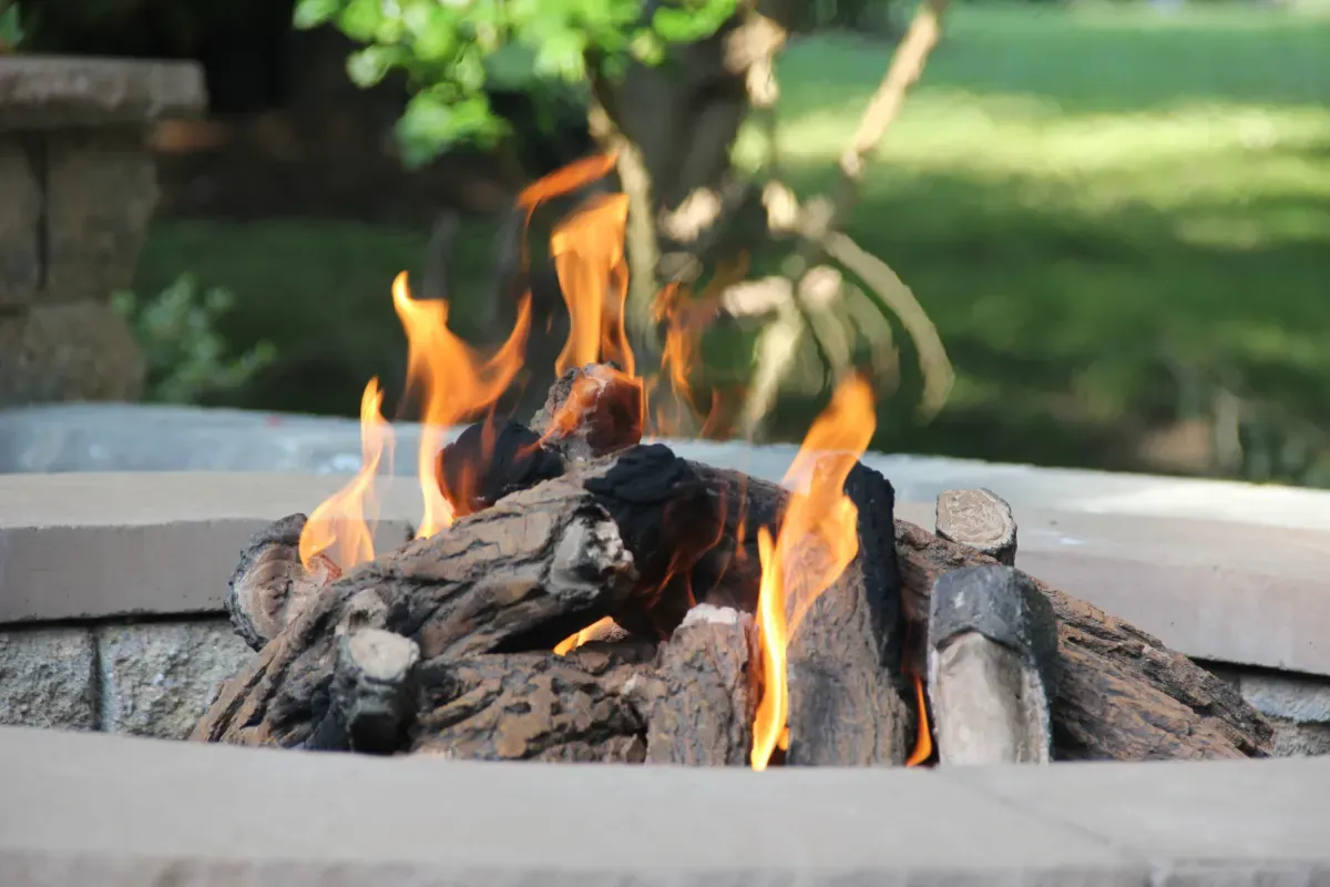 Fire burning in a built-in block fire pit with a smooth stone cap, with a green lawn behind it