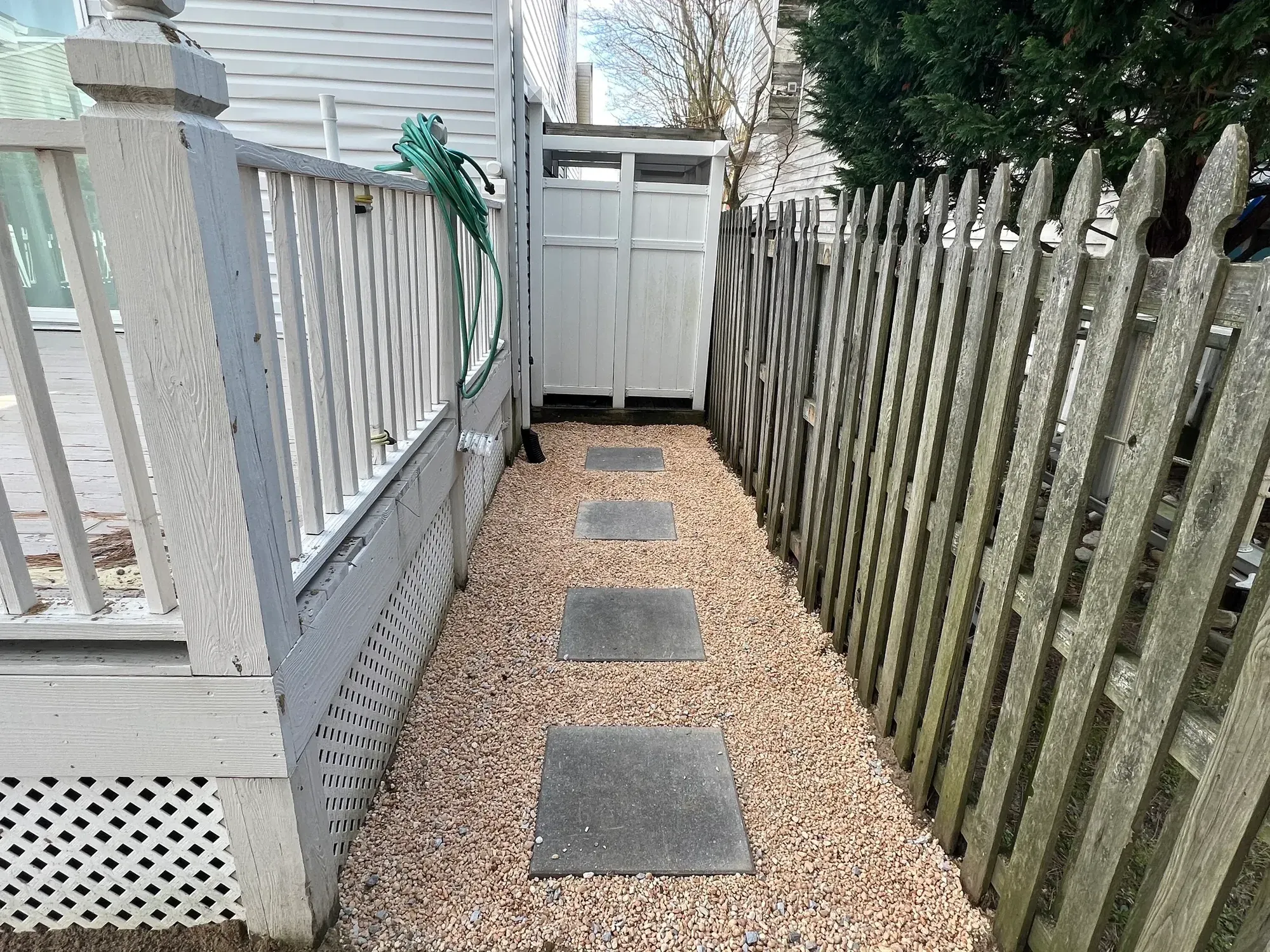 Narrow side yard finished with pea gravel and square stepping stones between a deck and a picket fence