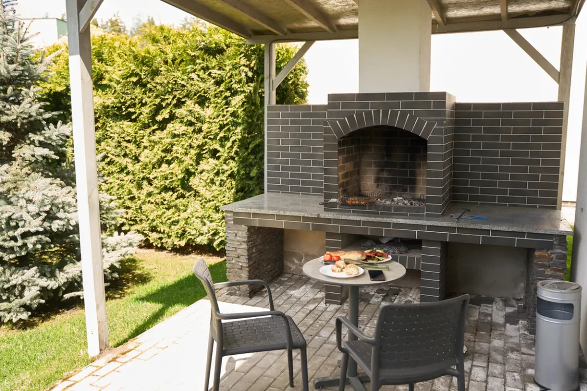 Built-in brick grill and stone counter on a paver patio under a covered pergola, with a small dining table