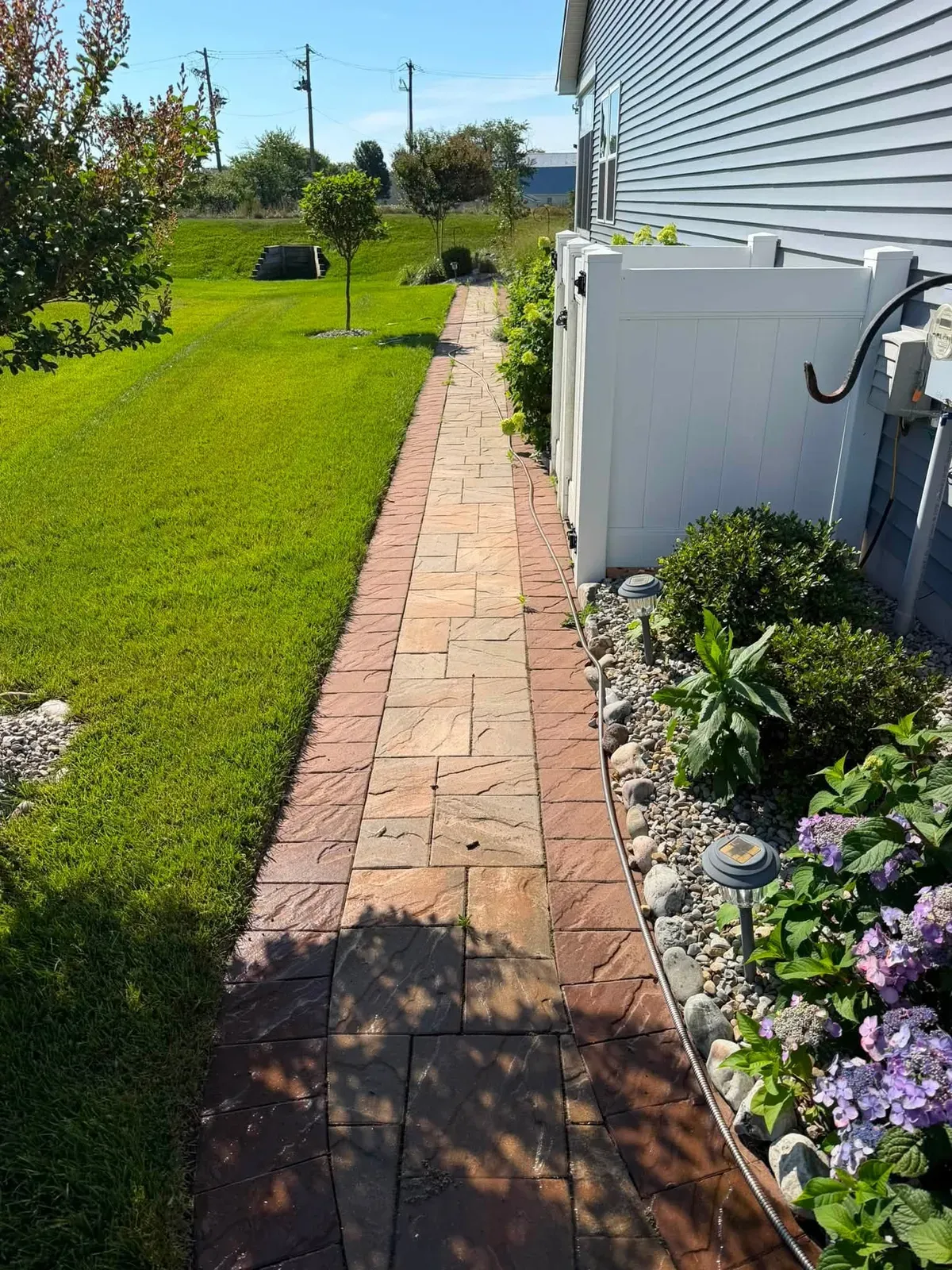 Long paver walkway with a dark border running between a lawn and a planted stone bed beside a house