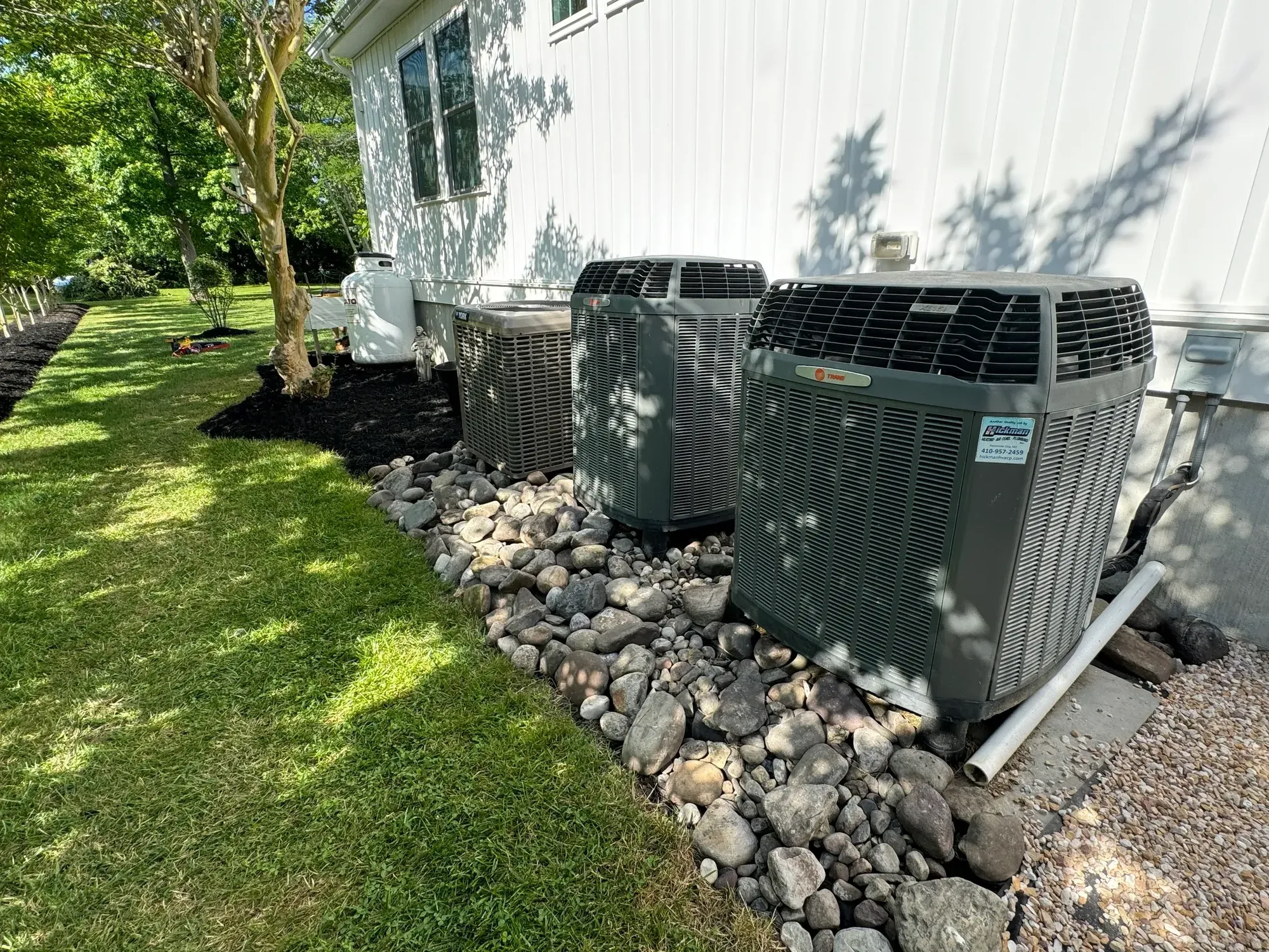 River rock bed around outdoor AC units, with a mulched bed and a small tree beside the house