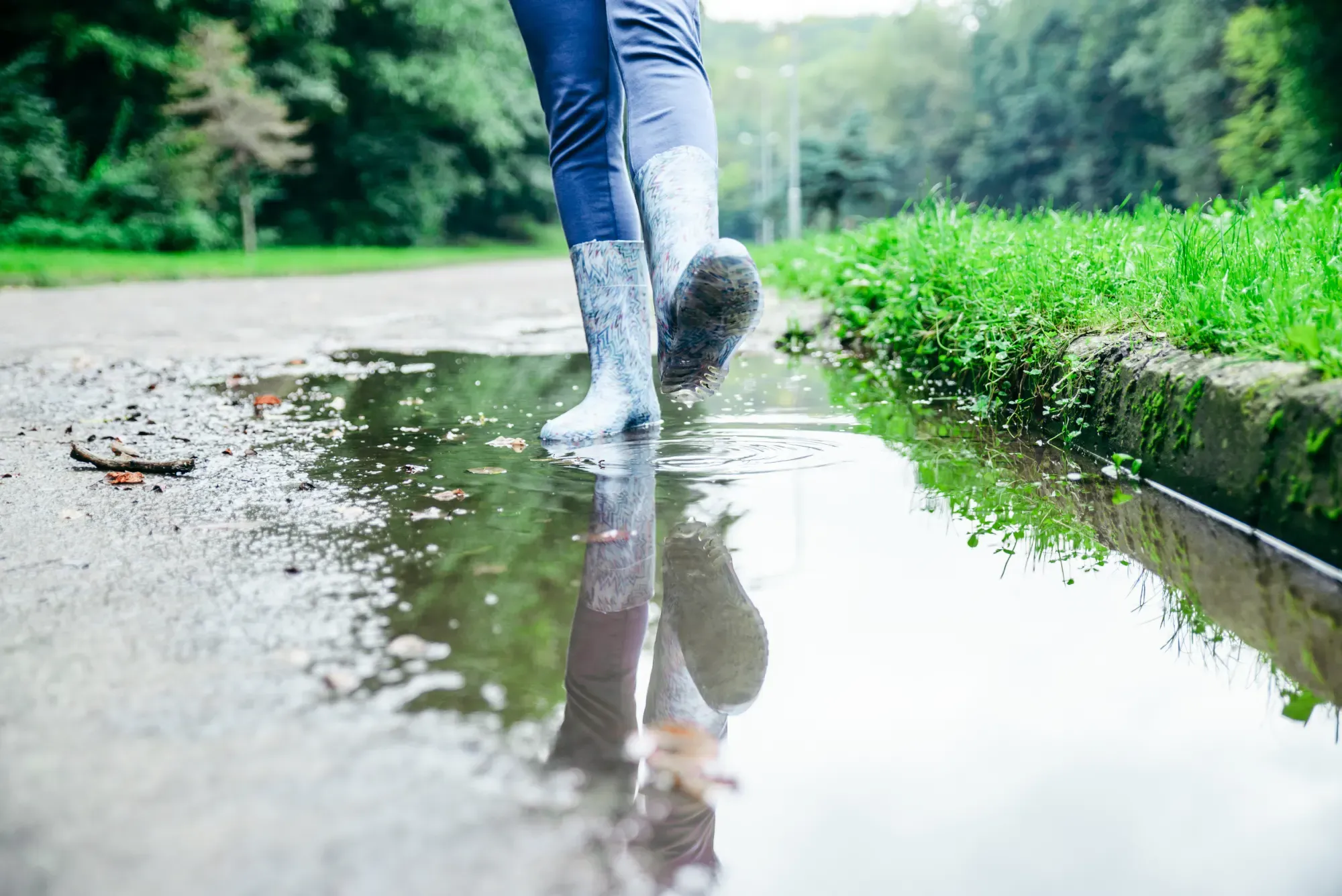 Rain boots splashing through a large puddle pooled along a grassy edge